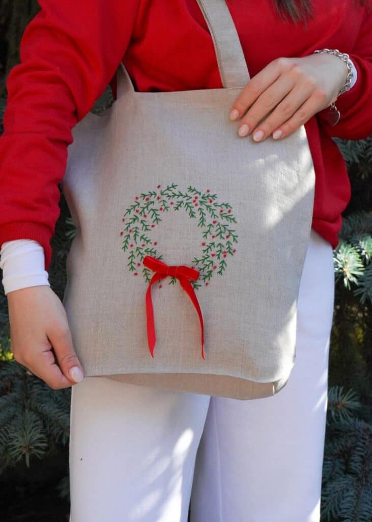 The Merry Carry - hand embroidered linen bag Woman holding a linen tote bag with a festive wreath and red bow design, wearing a red sweater and white pants outdoors. | Sky Rye Design Woman holding a linen tote bag with a festive wreath and red bow design, wearing a red sweater and white pants outdoors.