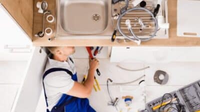 Plumber fixing a kitchen sink with tools and pipes around, showcasing home maintenance and repair work under the counter.