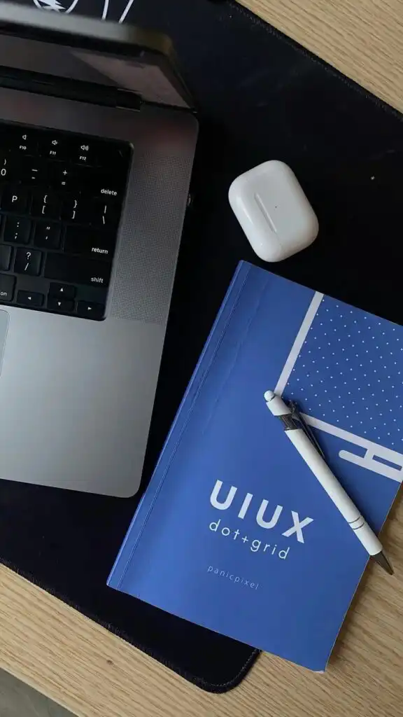 Laptop, AirPods, and a blue UI/UX notebook with pen on a wooden desk, ideal for digital design and productivity setups.
