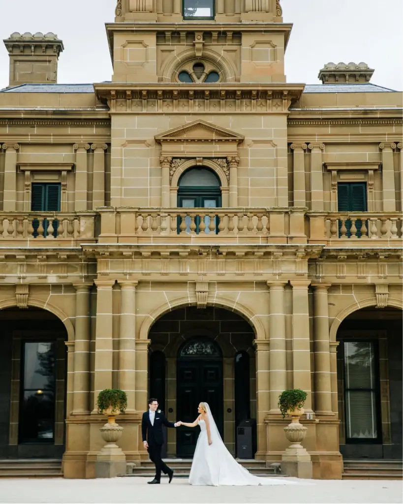 The-Werribee-Mansion-Wedding-Enquiry-Form_test400x500 Bride and groom holding hands in front of a historic stone building, creating a romantic and timeless wedding scene. | Sky Rye Design Bride and groom holding hands in front of a historic stone building, creating a romantic and timeless wedding scene.