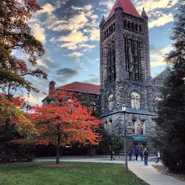Historic stone tower with red autumn tree at university campus against a blue sky, students walking by.