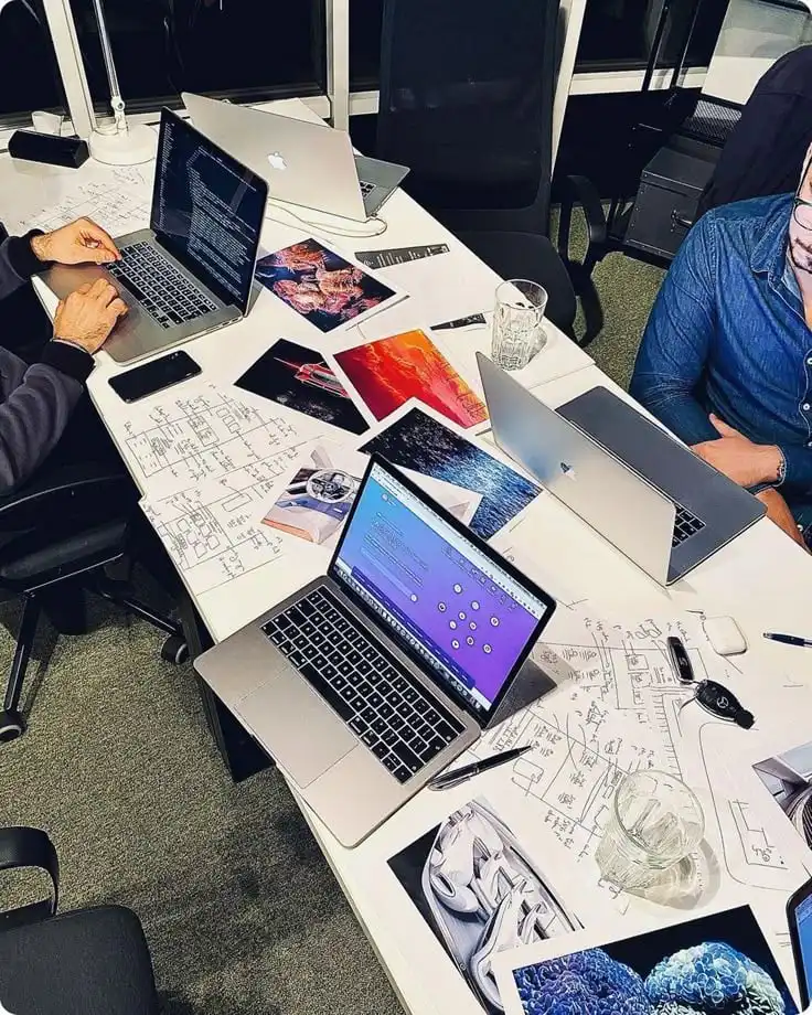 Team collaborating over laptops, planning with sketches and documents on a desk. Creative workspace with brainstorming in progress.