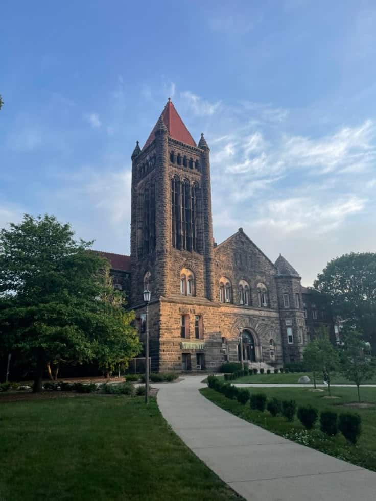 University of Illinois Historic stone building with a tall tower and red roof, surrounded by greenery under a clear blue sky. | Sky Rye Design Historic stone building with a tall tower and red roof, surrounded by greenery under a clear blue sky.