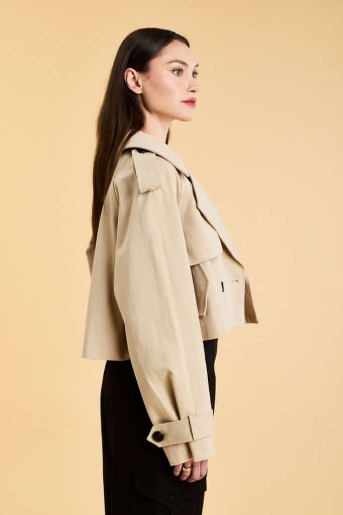 Victoria Cropped Trench Coat In Stock Now - XXS Profile of a woman in a beige trench coat with black pants standing against a beige background, showcasing classic autumn fashion. | Sky Rye Design Profile of a woman in a beige trench coat with black pants standing against a beige background, showcasing classic autumn fashion.