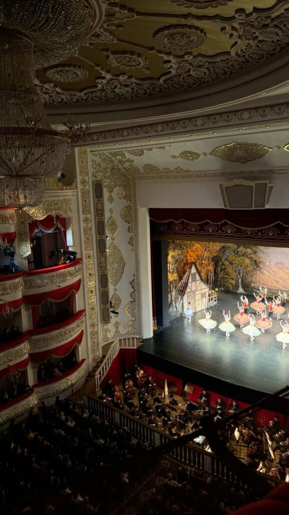 Vienna Opera House Ballet Elegant theater interior with ornate design, audience seated, and ballet dancers performing on stage, accompanied by an orchestra below. | Sky Rye Design Elegant theater interior with ornate design, audience seated, and ballet dancers performing on stage, accompanied by an orchestra below.