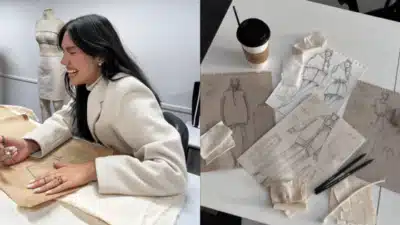 A fashion designer joyfully sketches on fabric, with garment designs and fabrics displayed on a nearby table with a coffee cup.