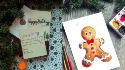 Holiday cards with festive greetings and a cheerful gingerbread man illustration, surrounded by paintbrushes and Christmas greenery.