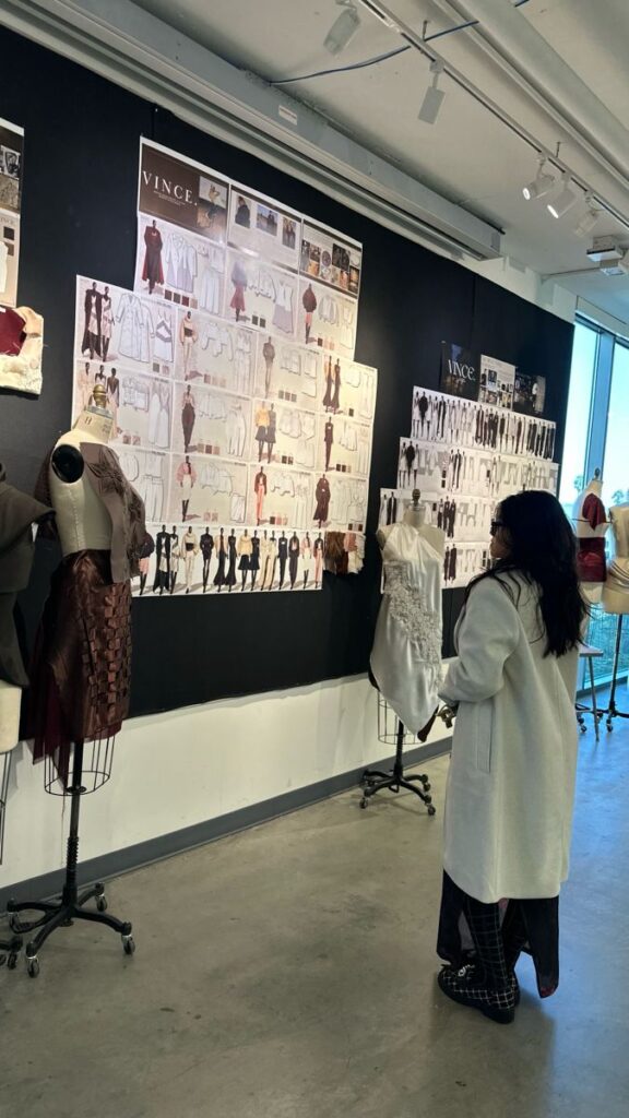 Fashion designer reviews clothing sketches and fabric samples on a wall display, next to mannequins in a bright studio.