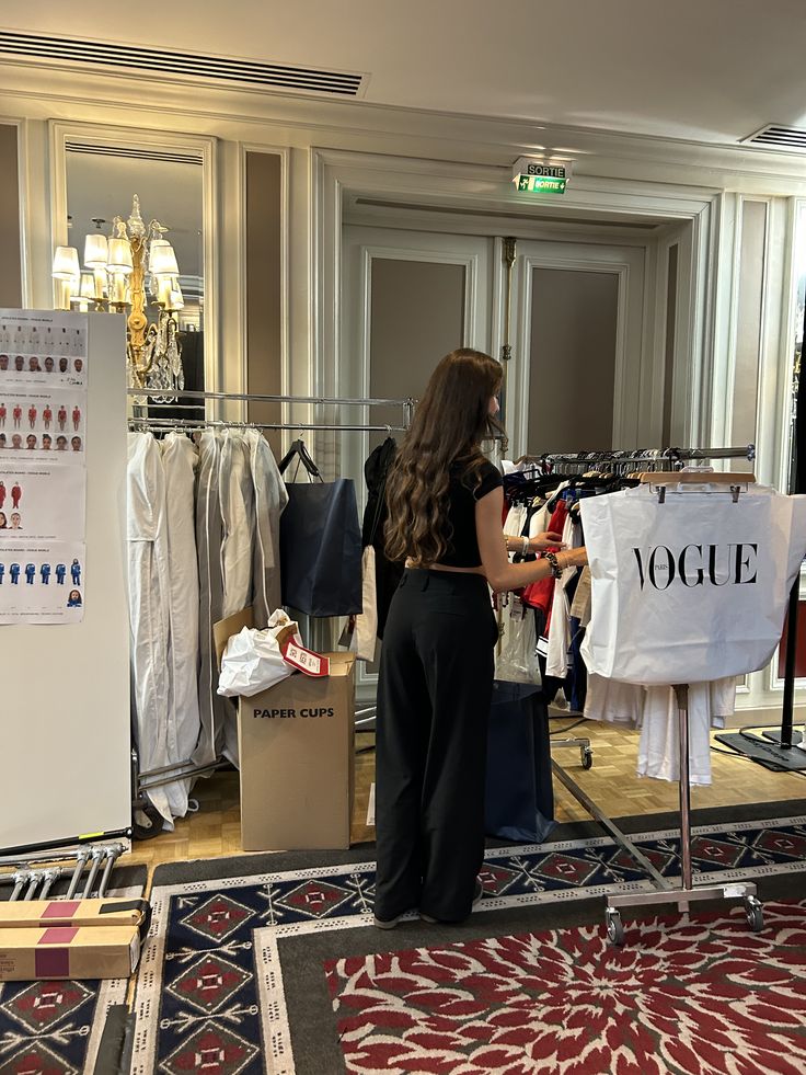 Person organizing clothes on a rack in a fashion showroom, with a Vogue bag and elegant decor, preparing for an event.