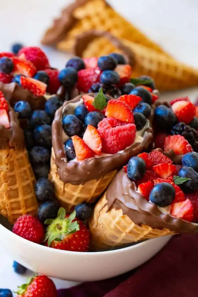 Chocolate-dipped waffle cones filled with fresh berries like strawberries, blueberries, and raspberries, served in a white bowl.