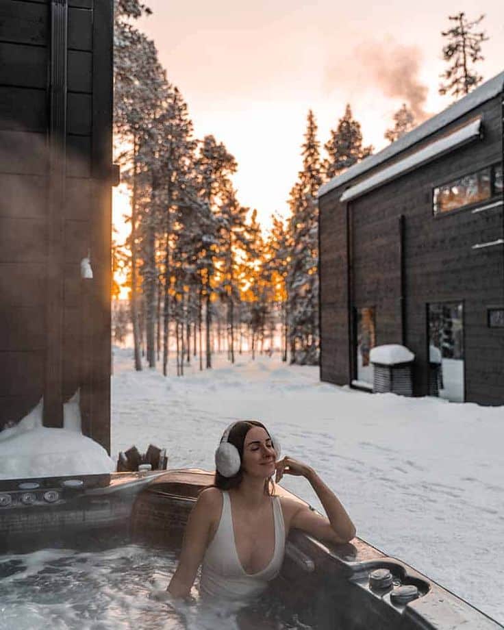 Woman relaxing in a hot tub with earmuffs in snowy landscape at sunset, next to wooden cabin in a winter forest setting.