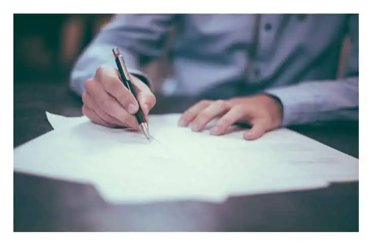 Person in a blue shirt writing on documents with a pen; close-up focused on hands and paperwork in an office setting.