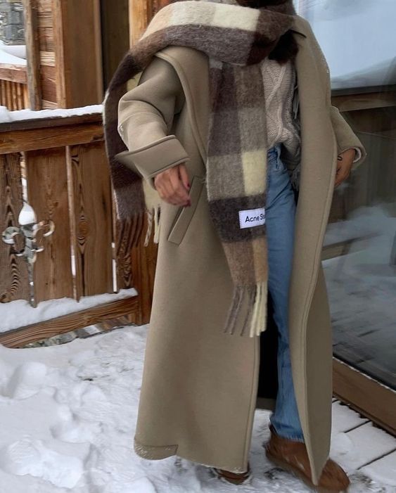 Winter Outfitz _ Acne Studios Person in a beige coat with a plaid scarf, jeans, and boots on a snowy porch, showcasing cozy winter fashion. | Sky Rye Design Person in a beige coat with a plaid scarf, jeans, and boots on a snowy porch, showcasing cozy winter fashion.