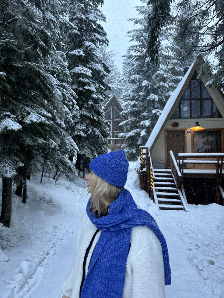 Winter Wonderland Woman in blue winter attire in snowy forest, standing near cozy A-frame cabin surrounded by snow-covered pine trees. | Sky Rye Design Woman in blue winter attire in snowy forest, standing near cozy A-frame cabin surrounded by snow-covered pine trees.