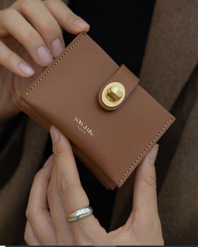 Brown leather wallet with gold clasp held in hands, featuring elegant stitching and a sleek design. Perfect for stylish everyday use.