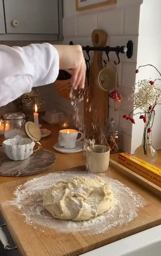 Cozy kitchen scene with dough on a floured board and a hand sprinkling flour. Lit candles and a rustic mug add warmth.