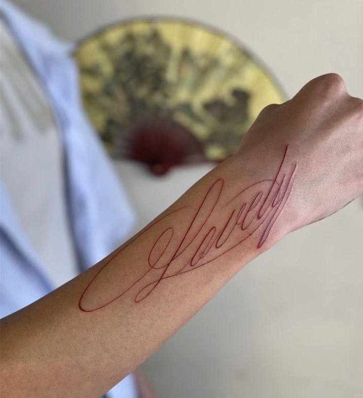 Arm tattoo with elegant cursive script reading Lovely in red, with a blurred fan in the background.