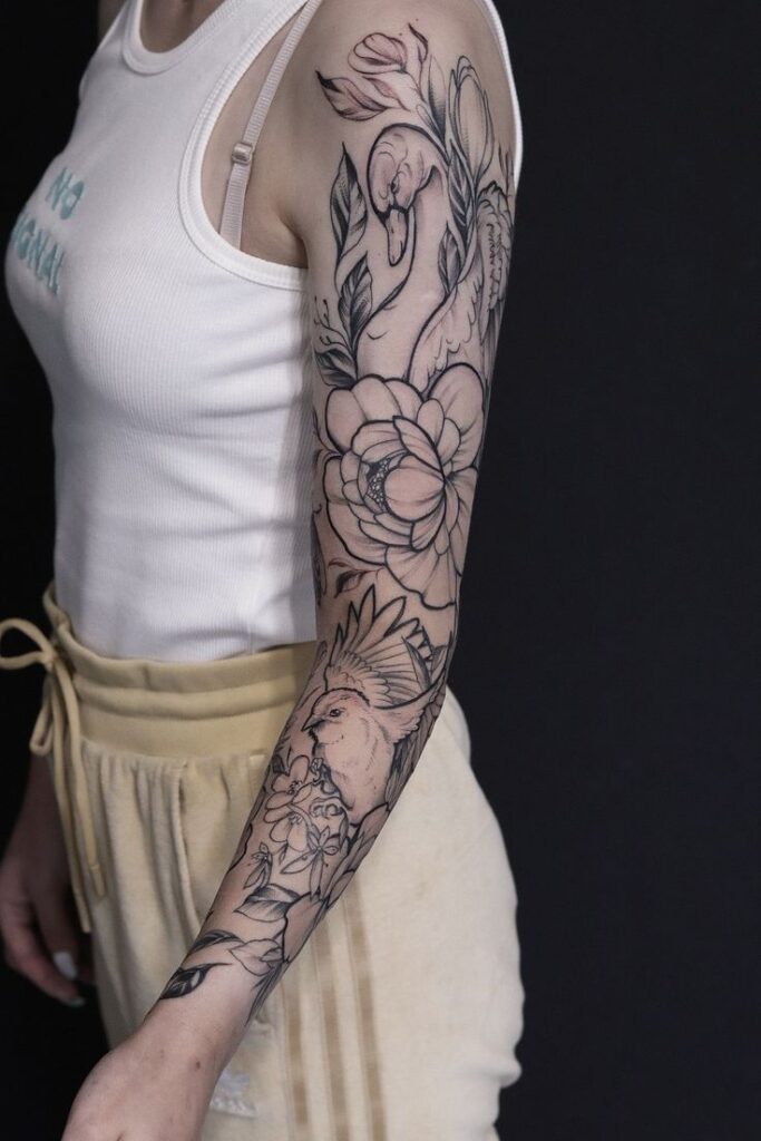 Arm sleeve tattoo featuring flamingo, floral, and bird designs on a woman wearing a white tank top and beige pants, black background.