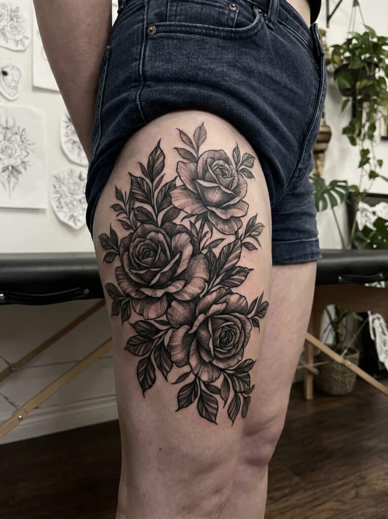 ai-photo-studio-1765823562840-2 Detailed black and gray rose tattoo on thigh with intricate leaf accents, tattoo sketches visible in background. | Sky Rye Design Detailed black and gray rose tattoo on thigh with intricate leaf accents, tattoo sketches visible in background.