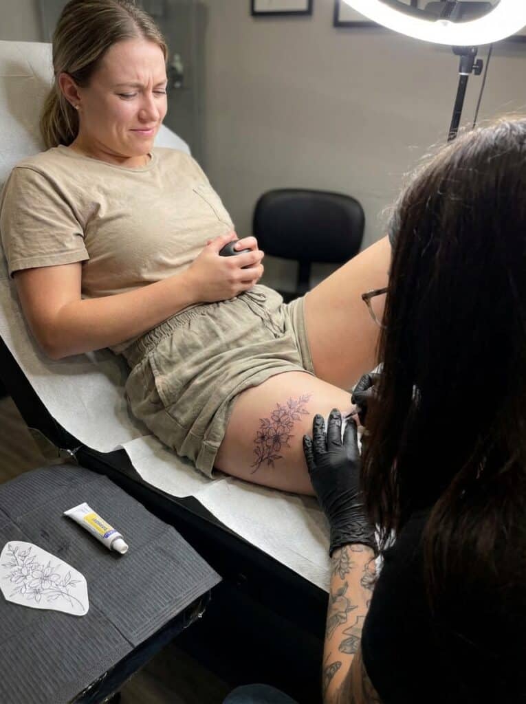 ai-photo-studio-1765904013086-1 Woman getting a floral tattoo on her thigh in a tattoo studio, showing expressions of nervousness and anticipation. | Sky Rye Design Woman getting a floral tattoo on her thigh in a tattoo studio, showing expressions of nervousness and anticipation.