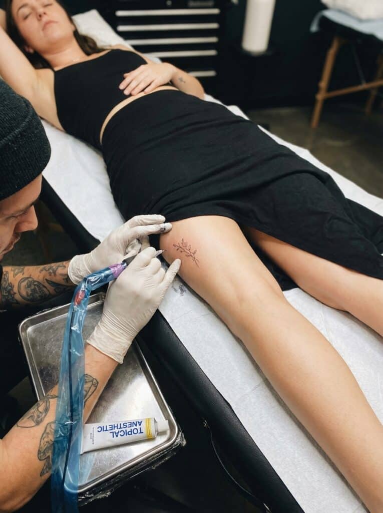 ai-photo-studio-1765904152055-1 Tattoo artist creating a floral design on a woman's thigh in a studio setting, highlighting tattoo artistry and body art. | Sky Rye Design Tattoo artist creating a floral design on a woman's thigh in a studio setting, highlighting tattoo artistry and body art.
