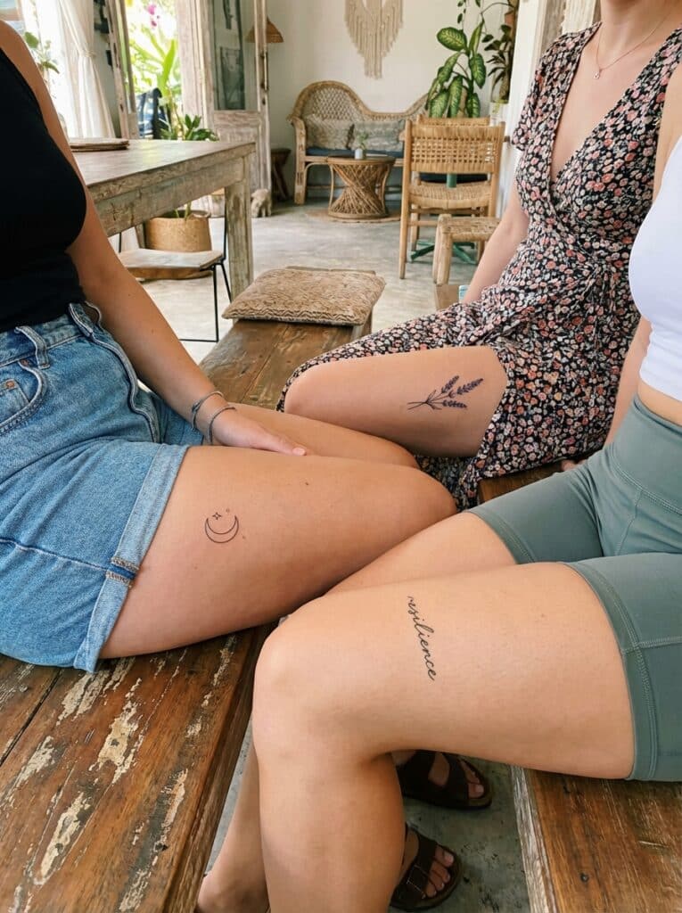 ai-photo-studio-1765904305107-1 Three women sitting, each displaying unique thigh tattoos: a moon, lavender, and the word resilience, in a cozy, rustic setting. | Sky Rye Design Three women sitting, each displaying unique thigh tattoos: a moon, lavender, and the word resilience, in a cozy, rustic setting. small thigh tattoos for women
