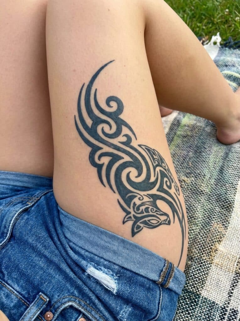 ai-photo-studio-1765905535970-1 Tribal tattoo on upper thigh with denim shorts, featuring intricate swirls and animal design, outdoors on a patterned blanket. | Sky Rye Design Tribal tattoo on upper thigh with denim shorts, featuring intricate swirls and animal design, outdoors on a patterned blanket.