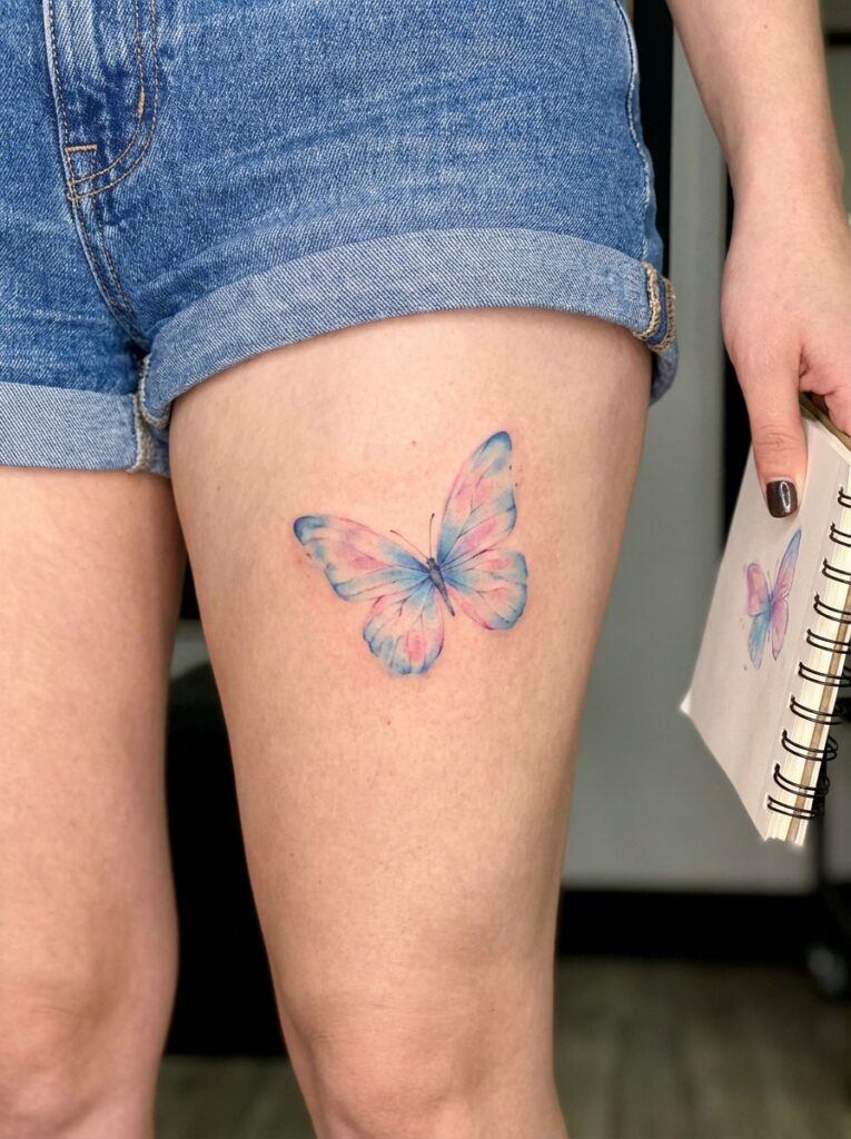 ai-photo-studio-1765905781076-2 Colorful butterfly tattoo on leg next to matching notebook sketch, showcasing vibrant blue and pink shades, in denim shorts. | Sky Rye Design Colorful butterfly tattoo on leg next to matching notebook sketch, showcasing vibrant blue and pink shades, in denim shorts.