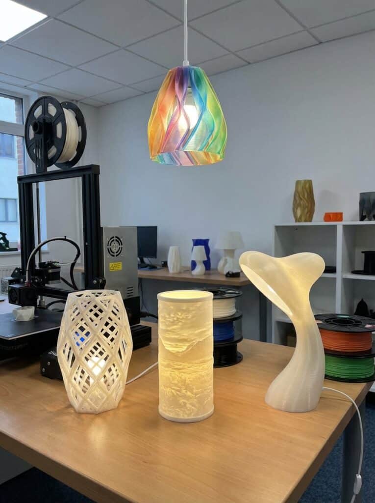 ai-photo-studio-1765921443856-2 3D-printed lamps in modern designs with a colorful pendant light, showcased in a tech workspace with 3D printer and filament spools. | Sky Rye Design 3D-printed lamps in modern designs with a colorful pendant light, showcased in a tech workspace with 3D printer and filament spools.