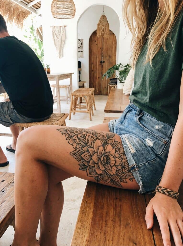 Woman with floral thigh tattoo sitting in a stylish, boho cafe wearing a green shirt and denim shorts, with natural decor elements.