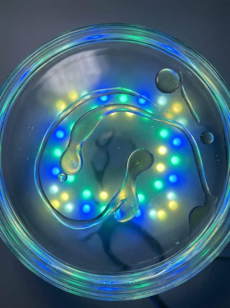 ai-photo-studio-1766413734162-1 Abstract close-up of colorful lights reflecting through clear liquid in glass dish; vibrant blue, green, and yellow hues. | Sky Rye Design Abstract close-up of colorful lights reflecting through clear liquid in glass dish; vibrant blue, green, and yellow hues.