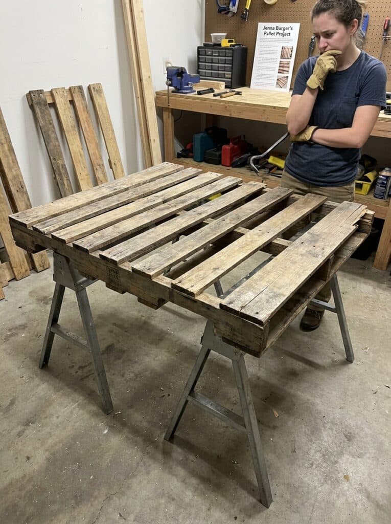 ai-photo-studio-1766515479737-2 Person pondering DIY wood pallet project in a workshop; table on sawhorses with tools. | Sky Rye Design Person pondering DIY wood pallet project in a workshop; table on sawhorses with tools.