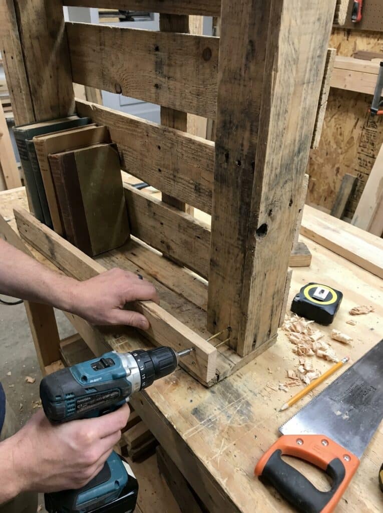 ai-photo-studio-1766515692104-2 Person using a drill to assemble a wooden pallet shelf in a workshop, with tools and books nearby. | Sky Rye Design Person using a drill to assemble a wooden pallet shelf in a workshop, with tools and books nearby.