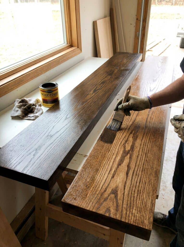 ai-photo-studio-1766515930284-2 Person applying wood stain to a wooden plank with a brush, highlighting DIY woodworking and home improvement projects near a sunny window. | Sky Rye Design Person applying wood stain to a wooden plank with a brush, highlighting DIY woodworking and home improvement projects near a sunny window.