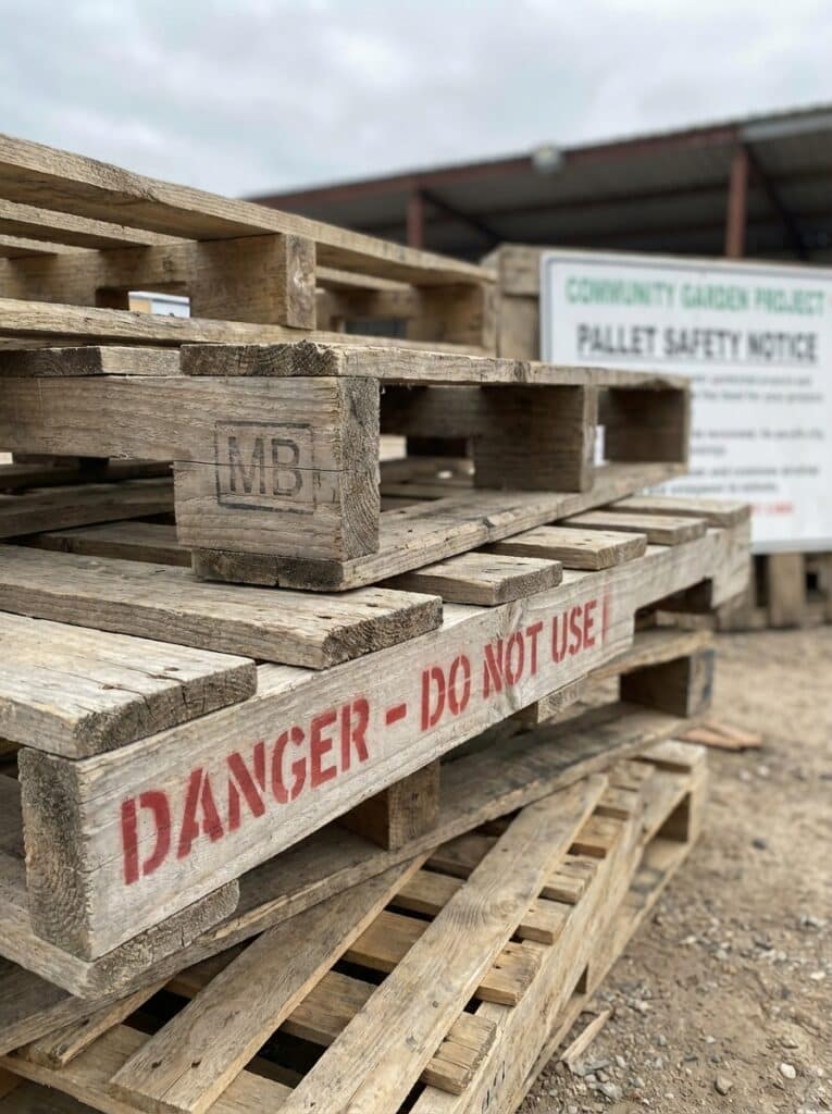 ai-photo-studio-1766516846141-2 Stack of wooden pallets marked Danger - Do Not Use, with a safety notice board in the background at a community garden project site. | Sky Rye Design Stack of wooden pallets marked Danger - Do Not Use, with a safety notice board in the background at a community garden project site.
