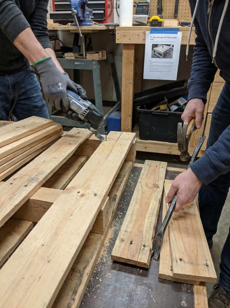 ai-photo-studio-1766673448535-2 Two people working on wooden pallets using a jigsaw and crowbar in a workshop setting. DIY wood project in progress. | Sky Rye Design Two people working on wooden pallets using a jigsaw and crowbar in a workshop setting. DIY wood project in progress.