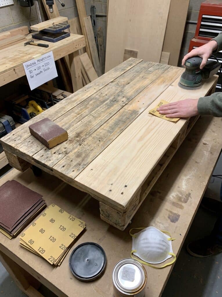 ai-photo-studio-1766673573716-1 Person sanding wooden pallet in a workshop with grit papers and tools, prepping for smooth finish. Woodworking DIY project. | Sky Rye Design Person sanding wooden pallet in a workshop with grit papers and tools, prepping for smooth finish. Woodworking DIY project.
