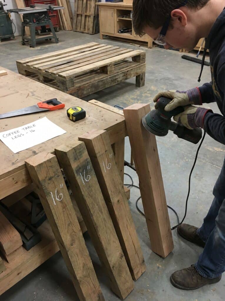 ai-photo-studio-1766673649814-2 Person sanding wooden coffee table legs in a workshop, surrounded by tools and a wooden pallet. | Sky Rye Design Person sanding wooden coffee table legs in a workshop, surrounded by tools and a wooden pallet.