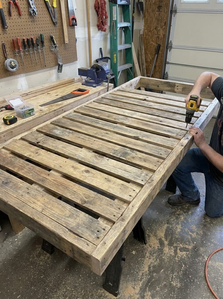 ai-photo-studio-1766673869558-2 Person building a wooden pallet bed frame with a drill in a workshop. Tools like saws and screwdrivers hang on the wall nearby. | Sky Rye Design Person building a wooden pallet bed frame with a drill in a workshop. Tools like saws and screwdrivers hang on the wall nearby.