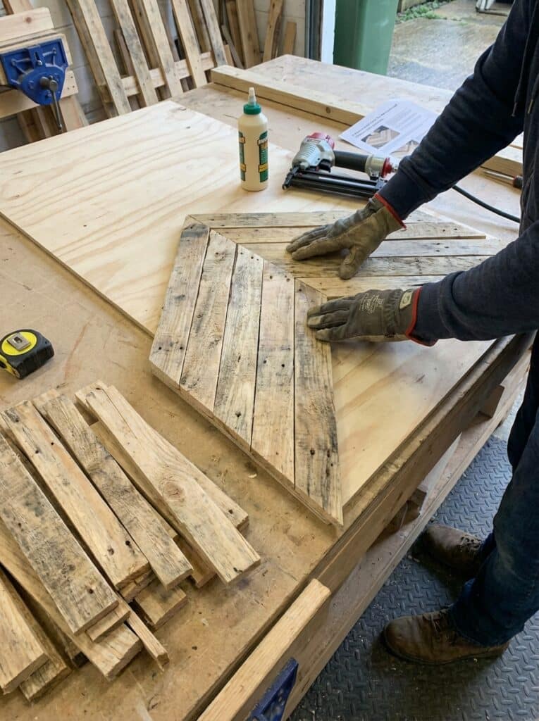 ai-photo-studio-1766673933381-2 Person crafting a wooden design with tools and glue in a workshop, emphasizing careful placement of reclaimed wood pieces. | Sky Rye Design Person crafting a wooden design with tools and glue in a workshop, emphasizing careful placement of reclaimed wood pieces.