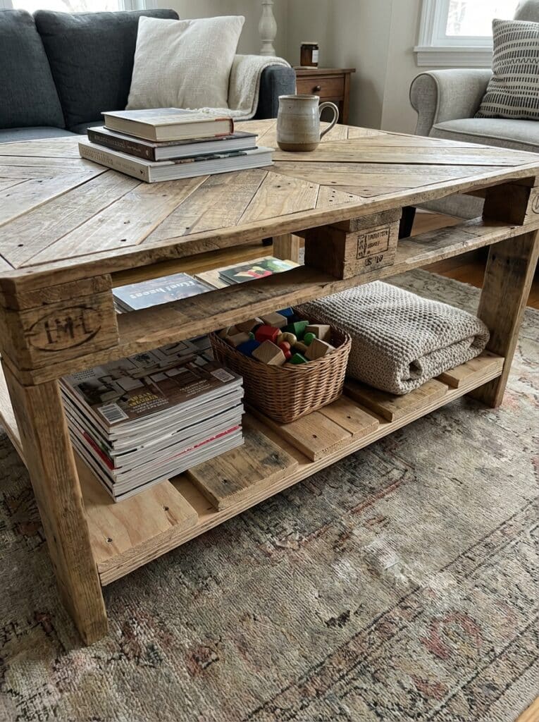 ai-photo-studio-1766674050850-2 Rustic wooden coffee table with books, a mug, magazines, a blanket, and a basket of toys, set in a cozy living room. | Sky Rye Design Rustic wooden coffee table with books, a mug, magazines, a blanket, and a basket of toys, set in a cozy living room.