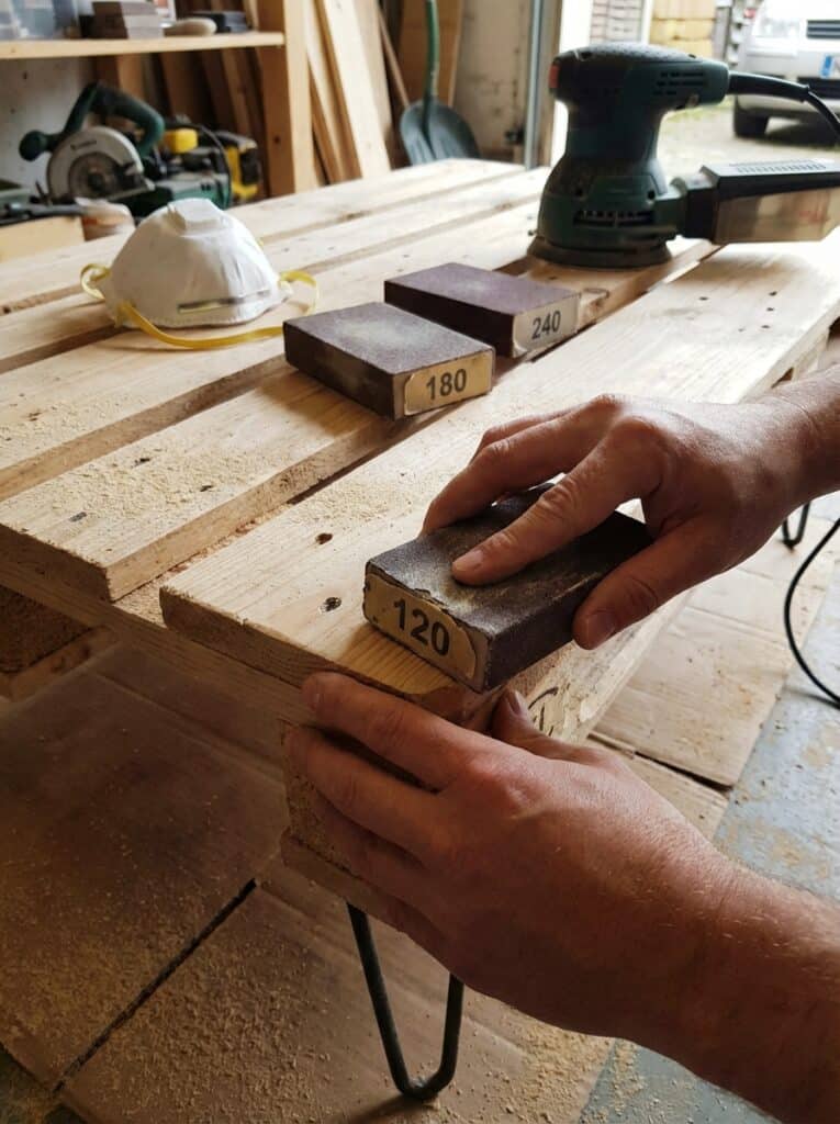 ai-photo-studio-1766674198977-1 Hands sanding a wooden pallet using 120-grit sandpaper, with tools and safety mask on the workbench. Woodworking in progress. | Sky Rye Design Hands sanding a wooden pallet using 120-grit sandpaper, with tools and safety mask on the workbench. Woodworking in progress.