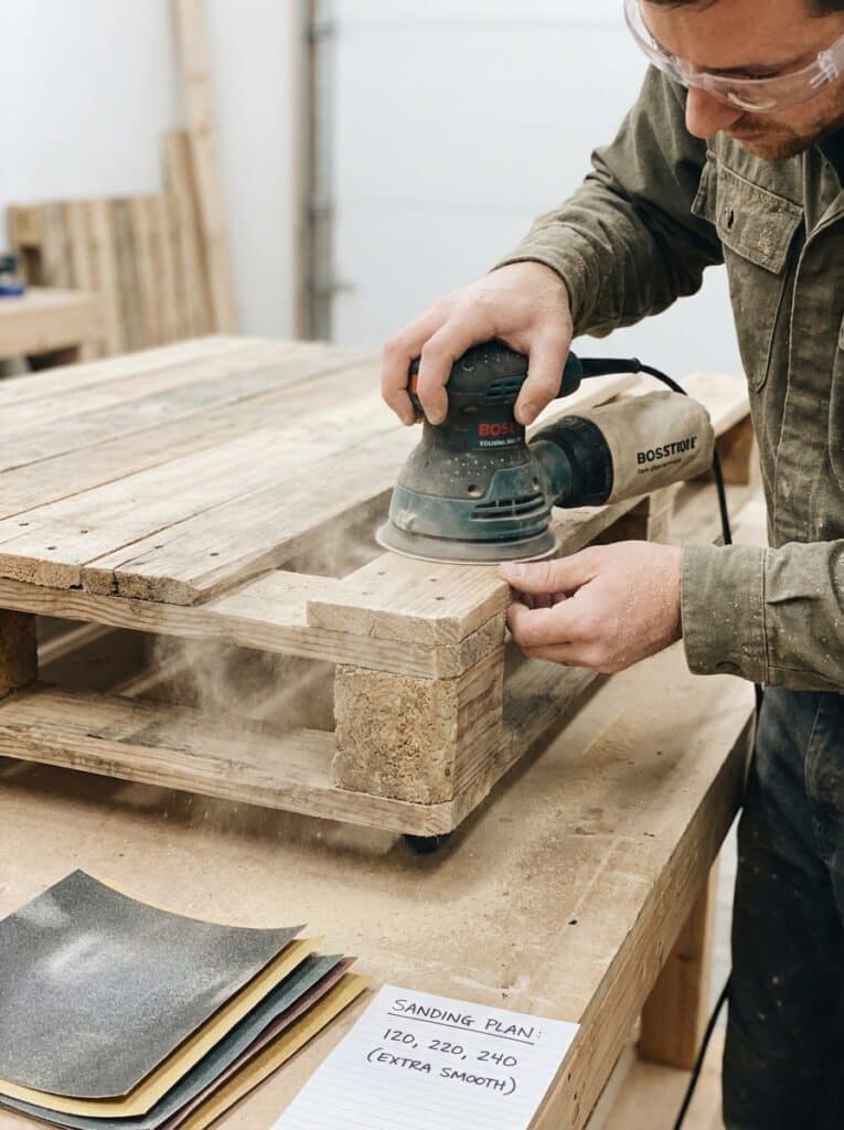 ai-photo-studio-1766674200277-2 Man sanding wooden pallet with an orbital sander in a workshop, following a sanding plan with various grit sandpapers on the table. | Sky Rye Design Man sanding wooden pallet with an orbital sander in a workshop, following a sanding plan with various grit sandpapers on the table.