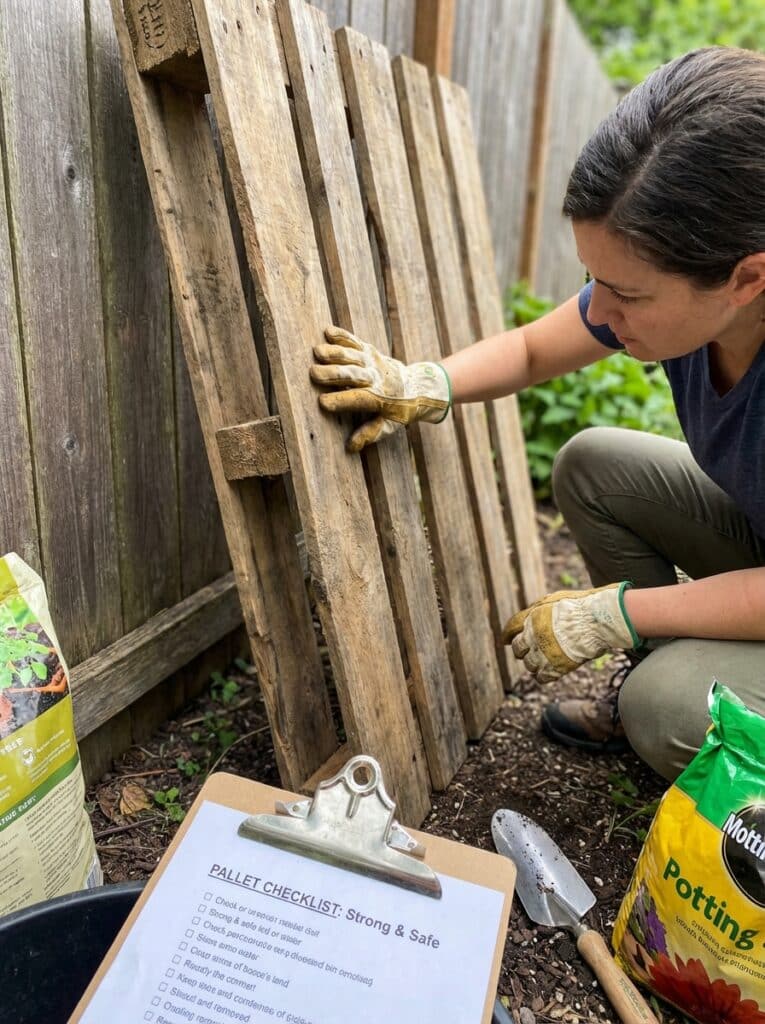 ai-photo-studio-1766675798108-2 Person examining wooden pallet in garden with gloves, checklist, and potting soil; preparing for a DIY project or garden improvement. | Sky Rye Design Person examining wooden pallet in garden with gloves, checklist, and potting soil; preparing for a DIY project or garden improvement.