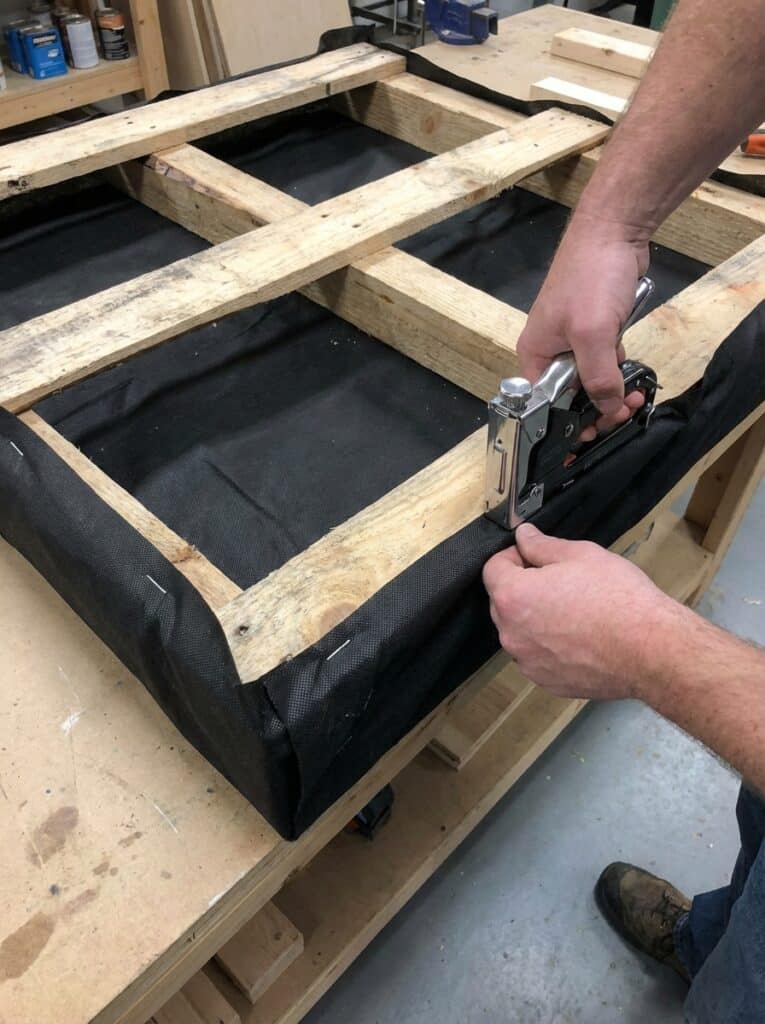 ai-photo-studio-1766678223702-1 Person using a staple gun to attach black fabric to a wooden frame in a workshop setting. | Sky Rye Design Person using a staple gun to attach black fabric to a wooden frame in a workshop setting.