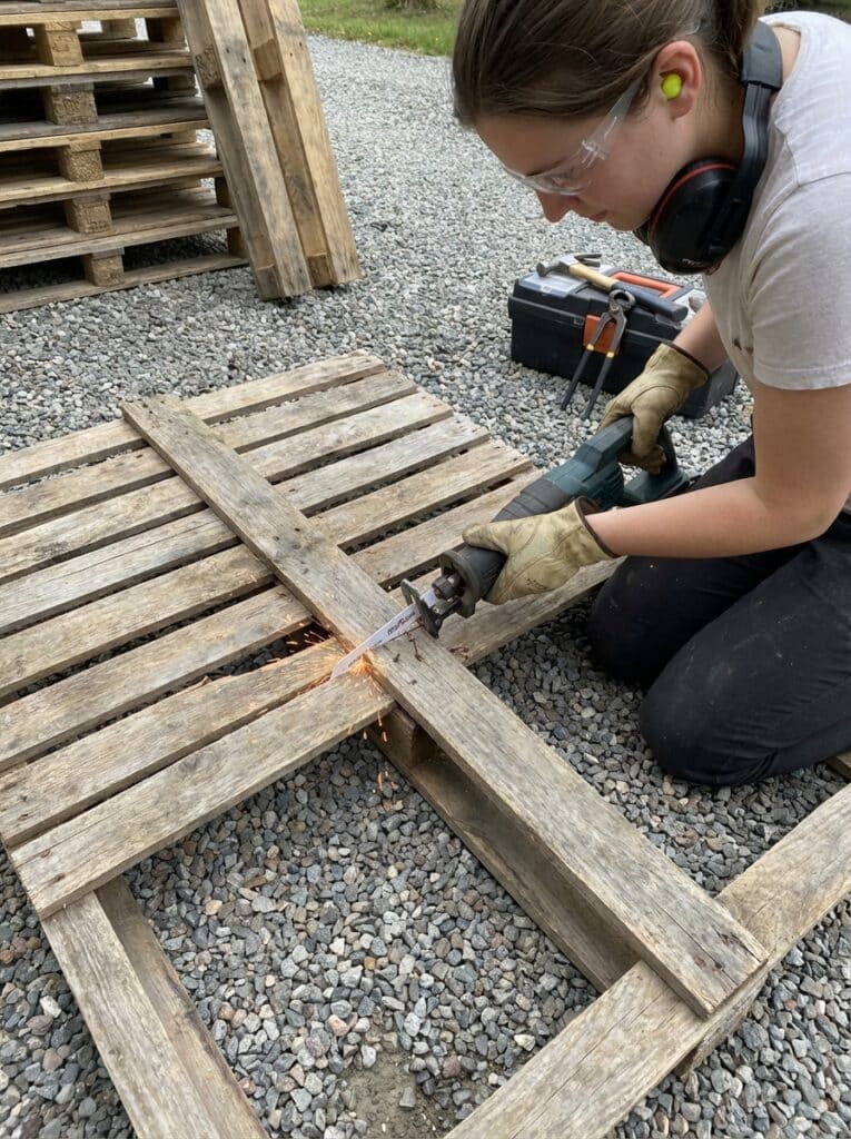 ai-photo-studio-1766940509844-1 Person using a reciprocating saw to cut a wooden pallet outdoors, wearing safety gear. | Sky Rye Design Person using a reciprocating saw to cut a wooden pallet outdoors, wearing safety gear.