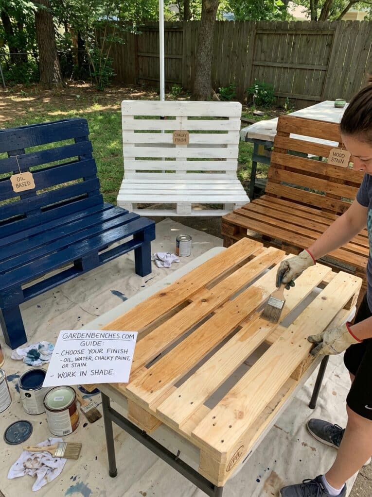 ai-photo-studio-1766940722858-1 Person painting a wooden pallet while other painted benches show oil, chalky, and wood stain finishes in a shaded backyard setting. | Sky Rye Design Person painting a wooden pallet while other painted benches show oil, chalky, and wood stain finishes in a shaded backyard setting.