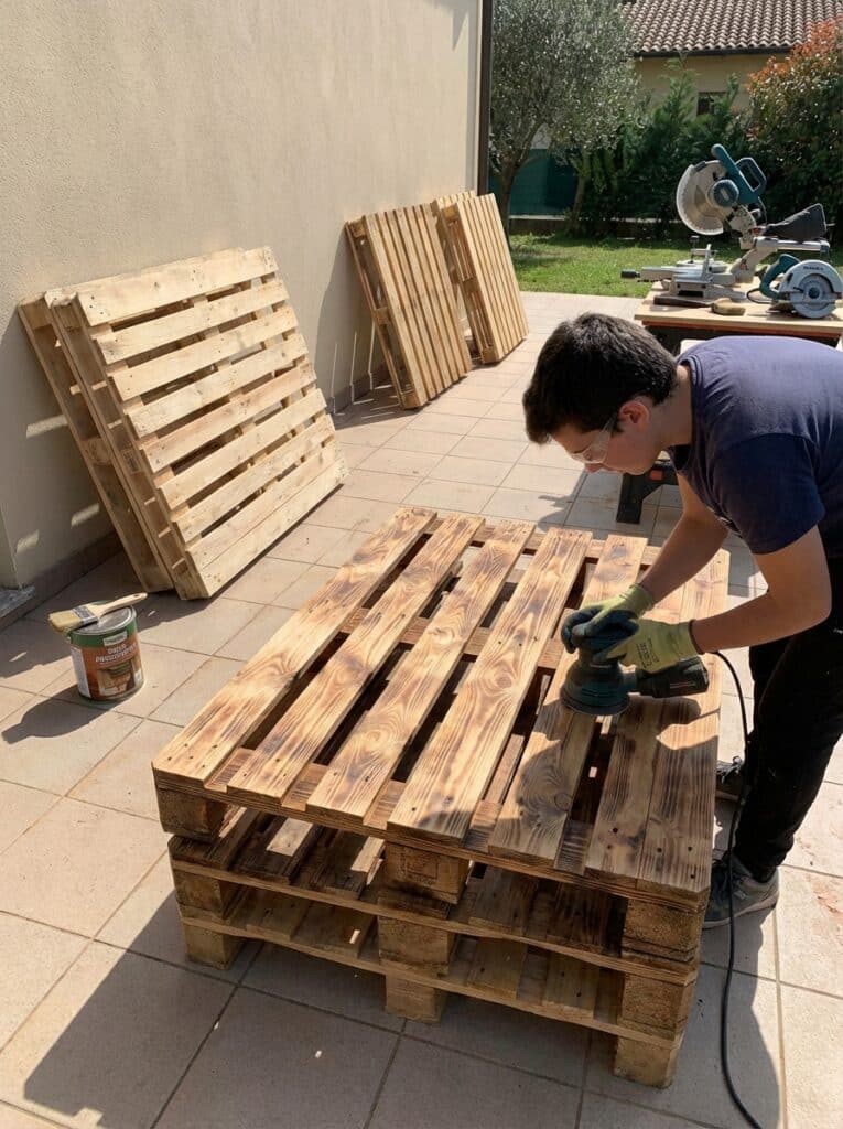 ai-photo-studio-1766940942134-2 Person sanding wooden pallets for a DIY project outdoors, surrounded by tools and paint, on a sunny day. | Sky Rye Design Person sanding wooden pallets for a DIY project outdoors, surrounded by tools and paint, on a sunny day.