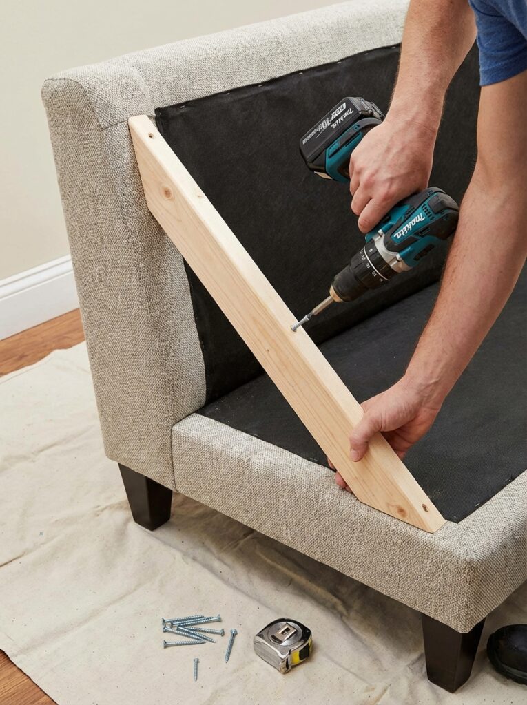ai-photo-studio-1766941082739-1 Person using a power drill to secure a wooden brace on a sofa, with screws and tape measure on a protective cloth nearby. | Sky Rye Design Person using a power drill to secure a wooden brace on a sofa, with screws and tape measure on a protective cloth nearby.