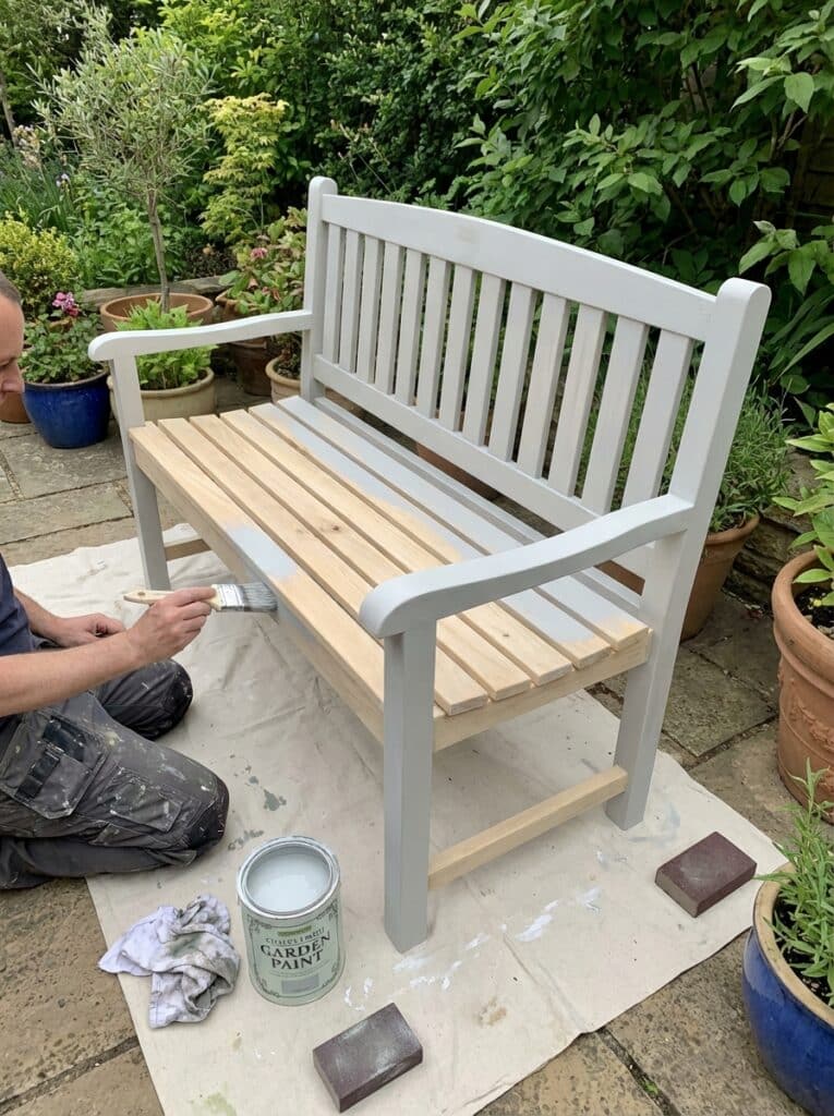 ai-photo-studio-1766941134357-2 Person painting a garden bench gray in a lush garden setting. Nearby are a paint can, brush, and sanding block on a tarp. | Sky Rye Design Person painting a garden bench gray in a lush garden setting. Nearby are a paint can, brush, and sanding block on a tarp.